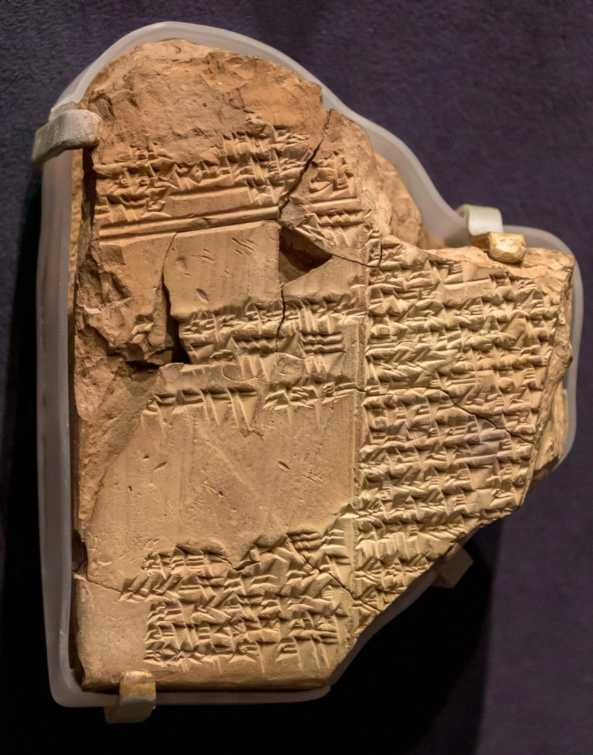 A broken clay tablet, sand-coloured and cracked, covered edge to edge in rows of small wedge-shaped cuneiform, held in clear mounts against a dark background.