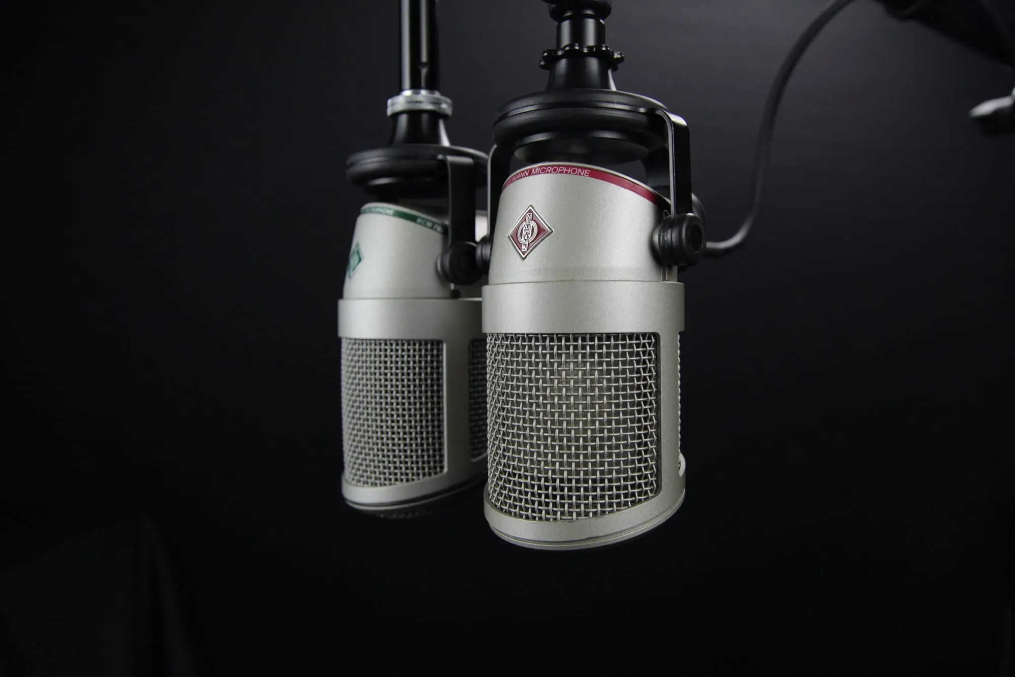 Two silver studio microphones on boom arms against a black background, one slightly behind the other, their mesh grilles facing the viewer.