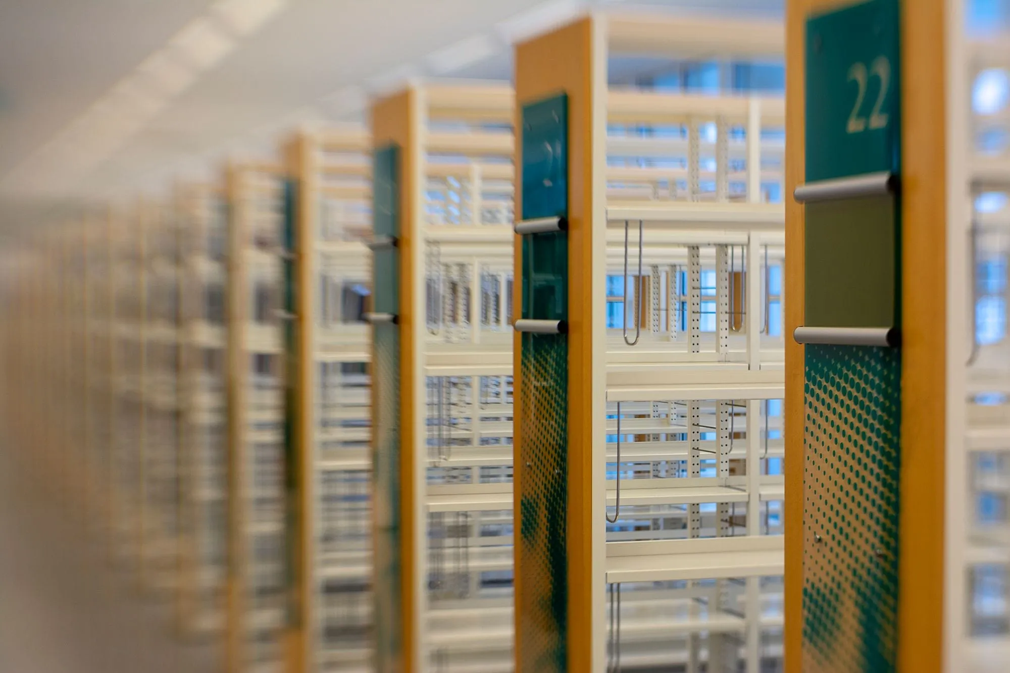 A row of empty library shelving units receding into soft blur, each with a pale wood end panel and a dark green sign; the nearest sign reads 22.