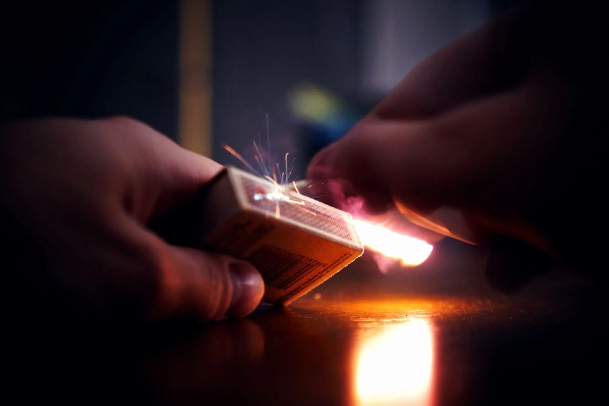 In near darkness, one hand strikes a match along a matchbox held in the other, throwing off sparks and a bright orange flare.
