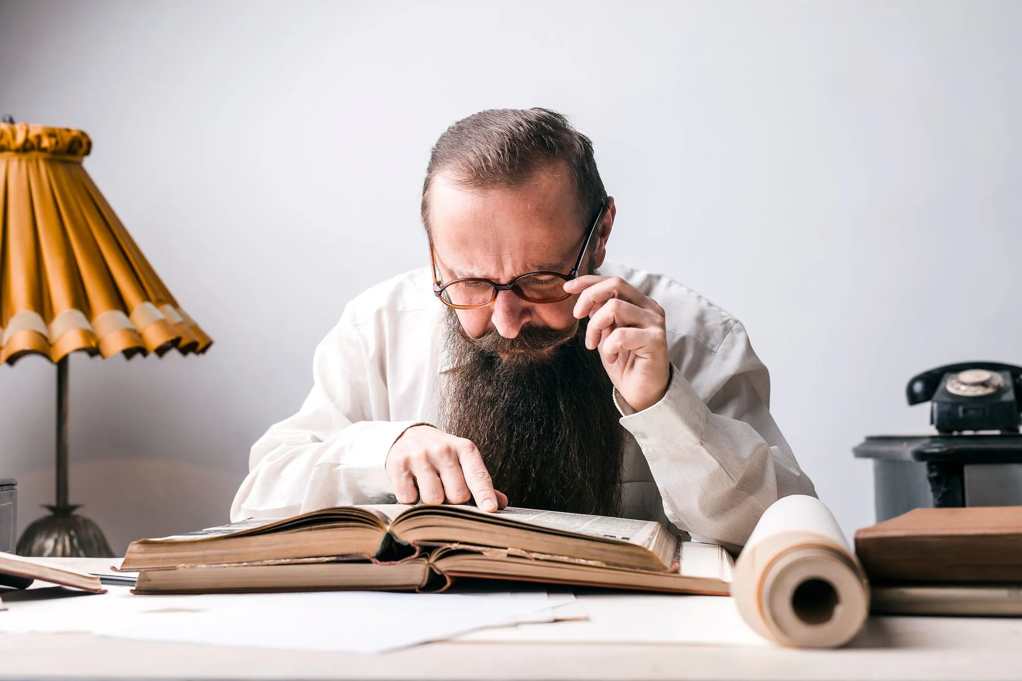 A man with a long dark beard, glasses and a white shirt leans over a thick, worn book open on a desk, one hand at his glasses and the other on the page, with a yellow pleated lamp and a black rotary telephone on either side.